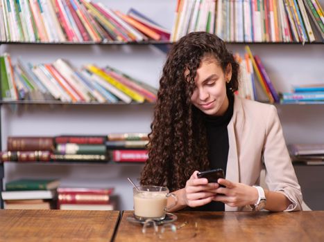 young woman checking her mobile phone while sitting with a hot drink. Shelf with books in the background