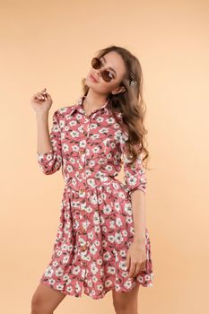 Studio portrait of stylish brunette young girl in sunglasses wearing nice summer dress with flower pattern and looking at camera. She is carrying a small white bag on chain and holding her skirt. Isolate.