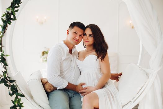 Beautiful couple of young people sitting on white swing with green leaves. Attractive woman and man looking at each other and holding hands together on female belly. Happy family waiting for baby.