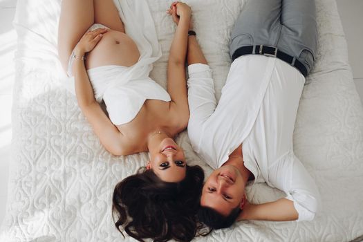 View from above of pregnant woman and caring husband lying on bed, looking at camera and posing. Lovely pair expecting little baby and touching big belly. Concept of pregnancy and awaiting.