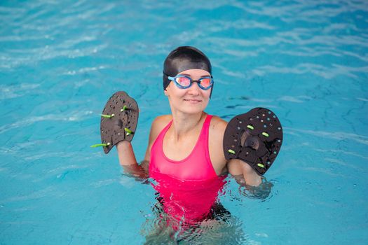 Young girl in goggles and cap swimming in blue water pool with special equipment for hand. Beautiful girl, wearing in pink swimsuit, standing in middle of water in pool, looking at camera and smiling,