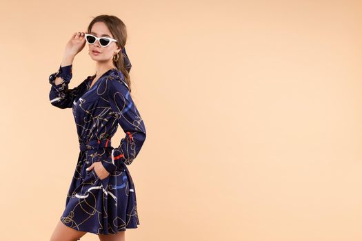 View from side of stylish woman in blue dress and glasses posing in studio. Young attractive girl wearing light clothes looking at camera on isolated background. Concept of fashion and trends.