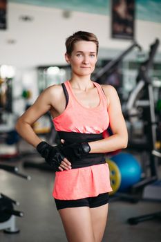 Serious sporty woman with strong body, wearing in pink t-shirt posing, looking at camera. Young athletic fit girl in gym, against simulators. Concept of sport bodybuilding.