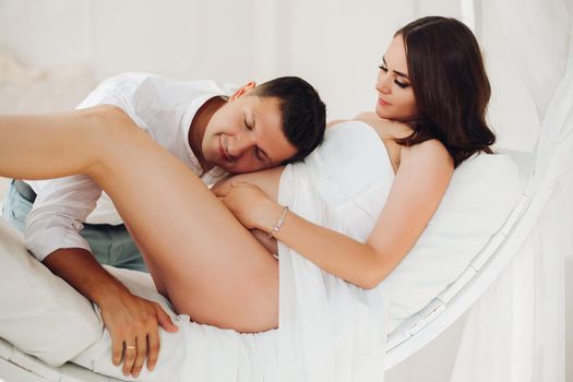 Happy family of young people waiting for baby. Beautiful brunette woman in white dress with naked leg lying on bed and looking at husband. Handsome man in casual clothes leaning to pregnant wife.