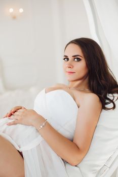 Beautiful brunette girl in white dress lying on bed and looking at camera. Portrait of gorgeous woman with make up and silver bracelet on hand. Happy young pregnant lady holding her hand on belly.