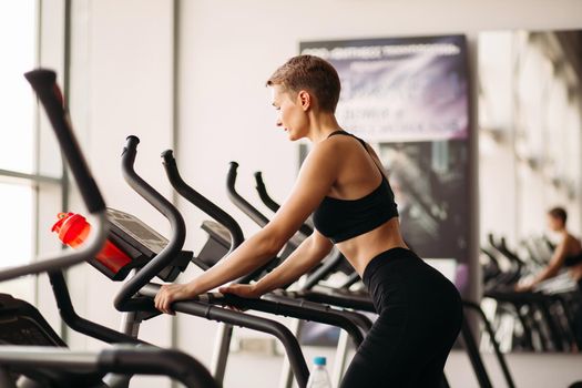Side view of fitness woman running on treadmill looking down, wearing in black sportswear. Healthy sporty woman doing cardio exercise on treadmill. Concept of sport and healthy lifestyle.