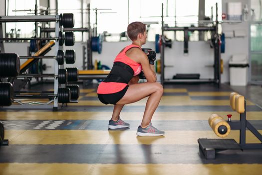 Back view of sporty woman wearing stylish sport wears, making exercise squatting with dumbbells in gym. Athletic and healthy girl with short hair working alone in gym. Simulators on background.