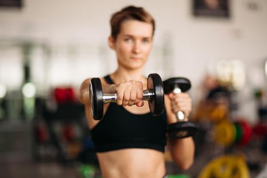 Selective focus of sporty woman wearing in black sportswear, lifting dumbbell weights in gym. Young attractive fitness girl showing her well trained body, developed muscles strong training.