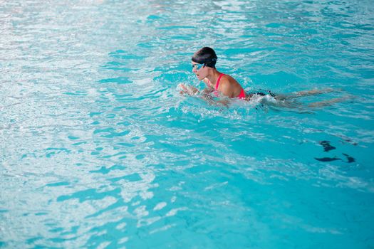 Side view of girl concentrated swimming forward, wearing in pink swimsuit, engaged in sports. Woman in goggles and cap swimming butterfly stroke style in the blue water pool. Sport lifestyle.