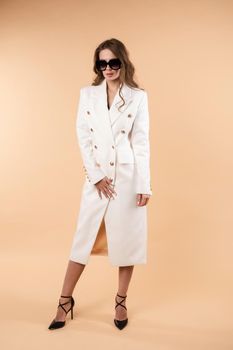 Elegant business woman in luxury clothes and shoes. Young stylish model in white and black look posing at camera. Gorgeous woman in white coat holding one hand on waist and in other one sunglasses.