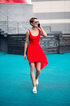 Young sexy woman wearing in red dress with deep neckline and sunglasses, talking on phone, walking outdoors. Hot girl with beautiful long legs wearing in white sandals on platform, looking sideways.