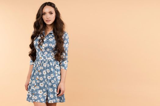Front view of pretty female in blue floral dress and sneakers looking at camera and smiling on isolated background. Charming brunette with long hair posing in studio. Concept of fashion and happiness.