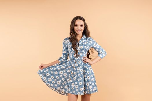 Studio portrait of positive brunette young woman with long brunette hair wearing trendy summer dress of blue color with flowers with skirt in her hand. She is smiling at camera. Isolate.