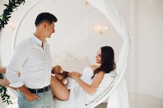 Side view of attractive pregnant woman lying on white bed and looking at beloved husband. Future parents touching belly with hands and expecting little baby. Concept of love and pregnancy.