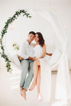 Beautiful couple of young people sitting on white swing with green leaves. Attractive woman and man looking at each other and holding hands together on female belly. Happy family waiting for baby.