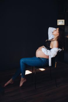 Side view of stylish pregnant woman in white shirt and jeans sitting in chair and posing. Attractive future mother awaiting little child and enjoying motherhood. Concept of beauty and pregnancy.