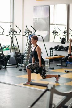 View of pretty healthy woman engaged in fitness in gym alone. Brunette woman with short hair, wearing in stylish sports wear, doing exercises with dumbbells, standing on special floor covering.