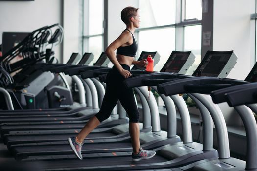 Side view of pretty fitness woman with short hairstyle running on treadmill at panoramic window, wearing in black sportswear. Healthy sporty woman doing cardio exercise on treadmill. Concept of sport.