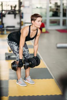 Brunette woman, wearing in stylish sports wear, perform physical exercises with dumbbells, standing on special floor covering. View of serious girl with short hairstyle working in fitness in gym.