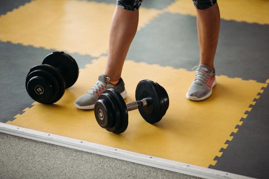 Close up of fitness equipment two dumbbell near female foot on yellow and black floor in modern gym. Concept of physical training and sport lifestyle.
