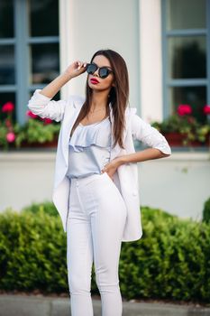 Portrait of a luxurious business woman with black glasses and on rebounds. The girl is dressed in a stylish white suit with a pale purple t-shirt. Isolated on the background of the building. Business woman concept.