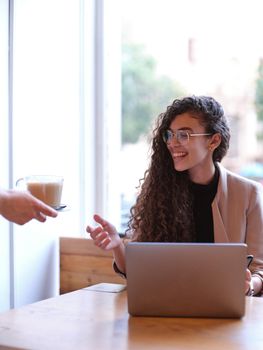 smiling woman receiving a coffee from the waiter while working on her computer, vertical background