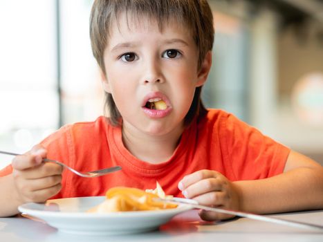 Kid in red t-shirt eats food with knife and fork. Fastidious boy with puzzled expression on face.