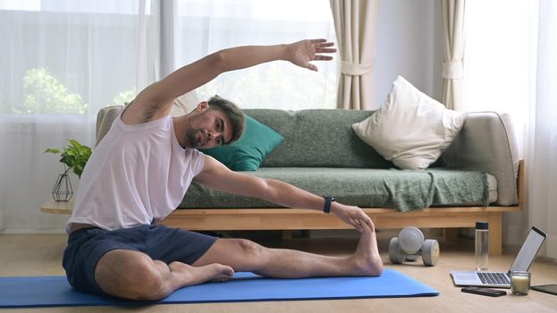 Young man in sports clothes is warming up, stretching while sitting on mat at home.