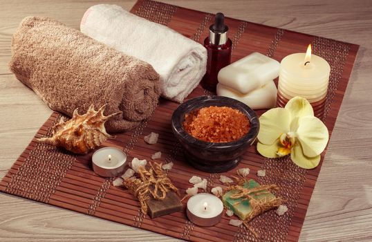 Spa and wellness setting with orchid flower, bowl with sea salt, seashell, bottle with aromatic oil, soap, candles and towels on brown bamboo napkin