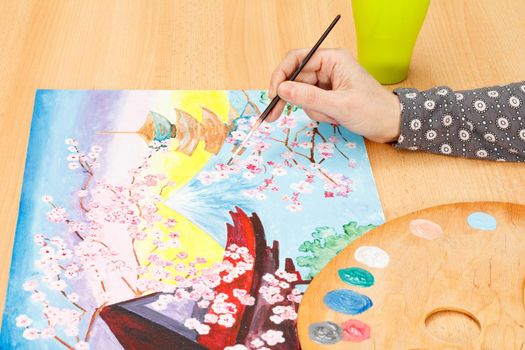 Woman's hand painting picturesque Japanese landscape indoors on the wooden desk with paints and paintbrush
