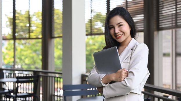 Pretty young female employee holding laptop and smiling to camera while standing on a terrace outside office.
