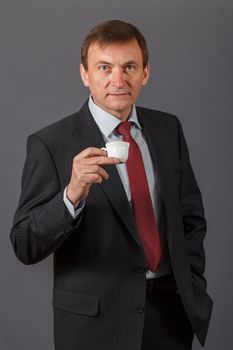 Confident and friendly elegant handsome mature businessman standing in front of a gray background in a studio taking a cup of coffee