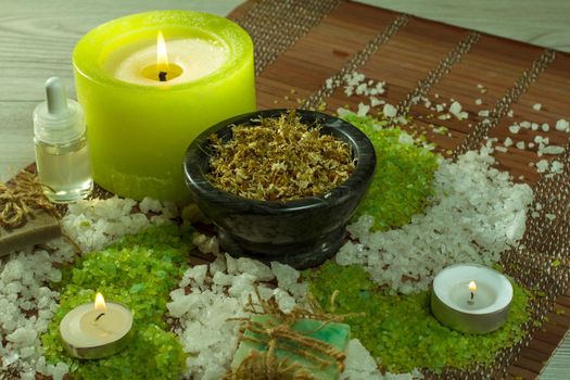 Spa accessories with soap, bowl with dried chamomile flowers, bottles with aromatic oil, sea salt, candles on bamboo napkin and wooden background