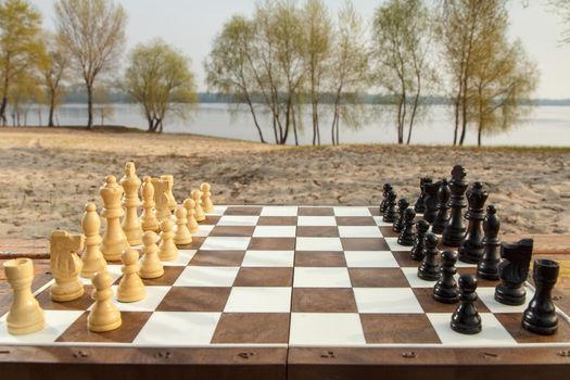 Chess board with chess pieces on river embankment background. Outdoors chess game with wooden chess pieces