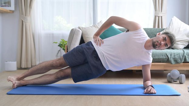 Young man doing side plank on blue mat in living room. Sport at home, workout, fitness and exercises.