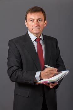 Confident and friendly elegant handsome mature businessman standing in front of a gray background holding a marker and writing in a notebook