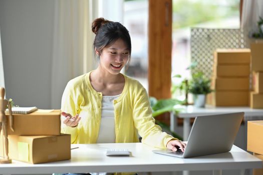 Smiling young female entrepreneur checking online order on digital tablet and preparing parcel boxes for delivery to customers.