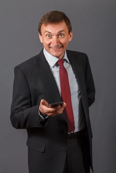 Elegant handsome mature businessman standing in front of a gray background in a studio wearing a nice suit holding a sell phone and having funny facial expression.