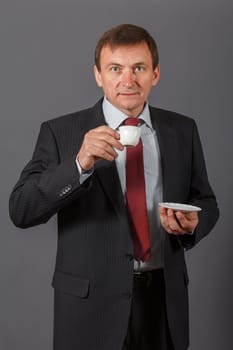 Confident and friendly elegant handsome mature businessman standing in front of a gray background in a studio taking a cup of coffee