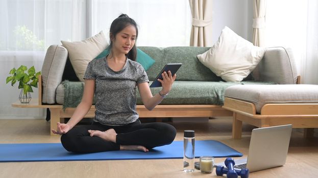 Beautiful young woman sitting in yoga mat and watching fitness lessons online on digital tablet.