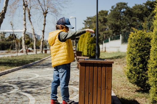 Schoolboy kid throwing the trash into dumpster. Boy using recycling bin to throw away the litter. Caucasian child recycles the junk into the trash-can..