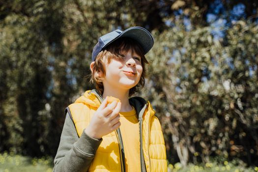 Smiling cheerful child kid in yellow vest and green hoodie eats crisp snacks outdoors in public park. Schoolboy boy enjoying consumes chews junk food outside with trees vegetation on the background.