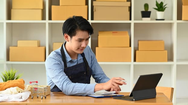 Young Asian man checking online order on digital tablet and preparing parcel boxes for delivery to customers. E-commerce, Online selling concept.