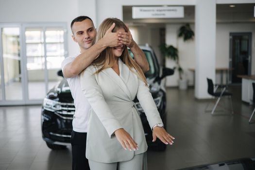 Man close eyes to his wife and makes a surprise buying car. Man and woman in car showroom. Happy young couple.