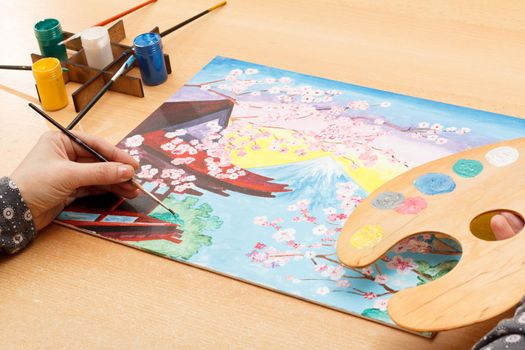 Artist painting picturesque Japanese landscape indoors on the wooden desk with palette, paints and paintbrushes