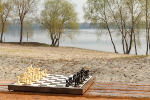 Chess board with chess pieces on wooden bench with river embankment background. Outdoors chess game with wooden chess pieces