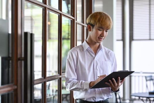 Asian male office worker reading information on clipboard while standing at modern office.