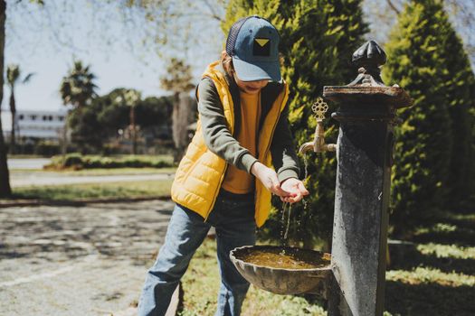 Kid schoolboy in casual clothing washing his hands in the street old fashioned drinking water fountain. Boy in blue jeans and yellow vest playing with water from drinking sprinkler in city park.