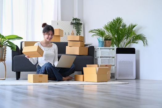 Happy female online seller checking online order on laptop and preparing packages for delivery to customers.