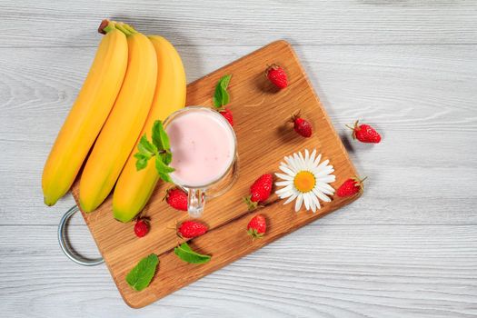 Glass of delicious milk shake with mint and fresh strawberries, banana, chamomile on a wood cutting board Top view